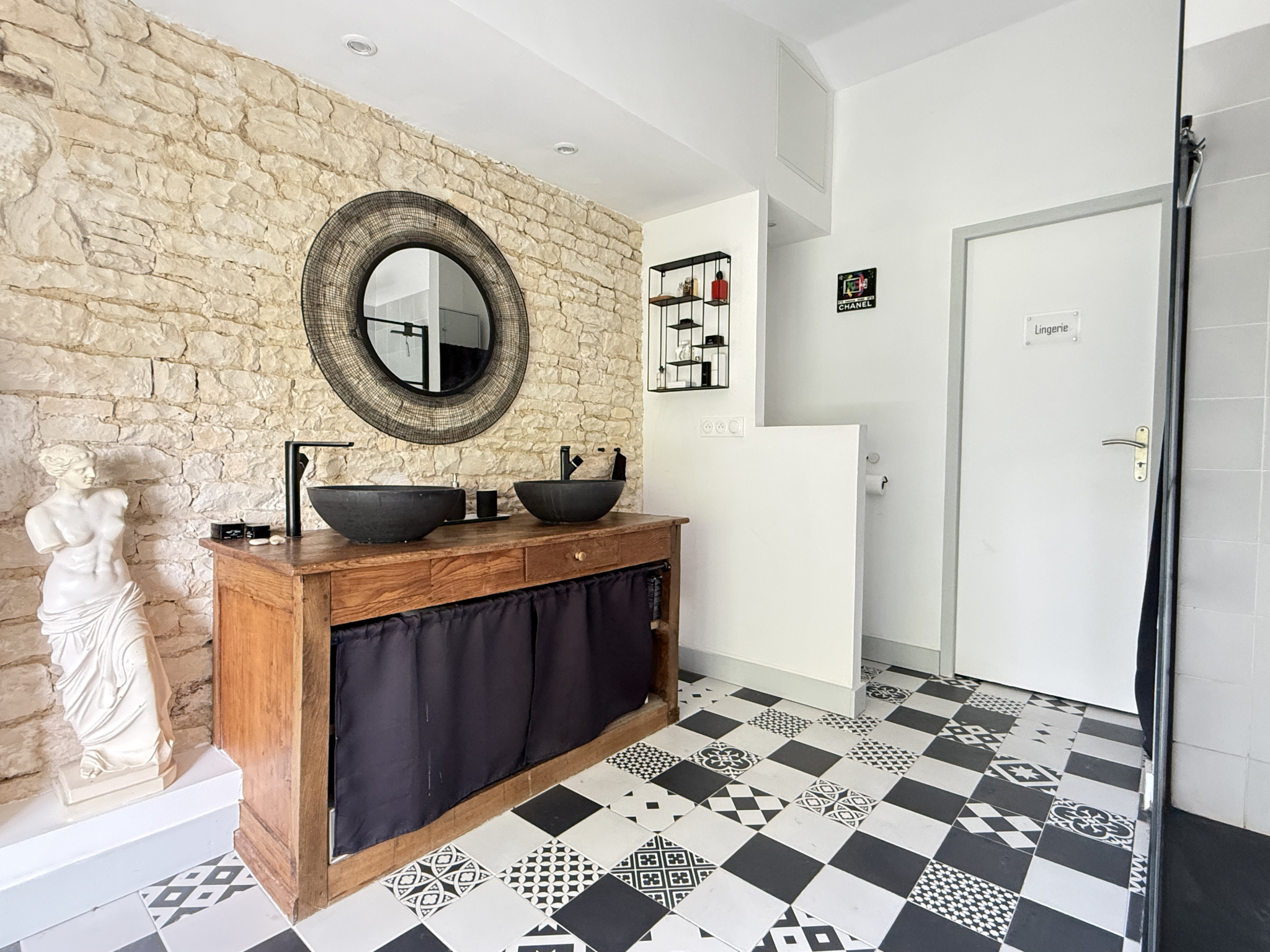 Salle de bain rénovée avec murs en pierre, lavabo en bois et carrelage noir et blanc.