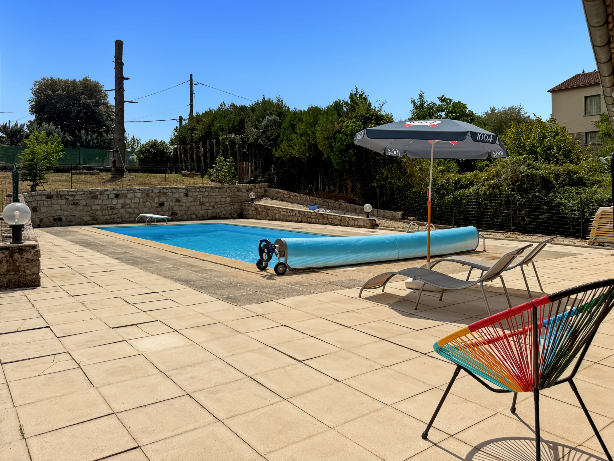 Piscine avec transats et parasol dans un jardin arboré, maison en pierre en arrière-plan.
