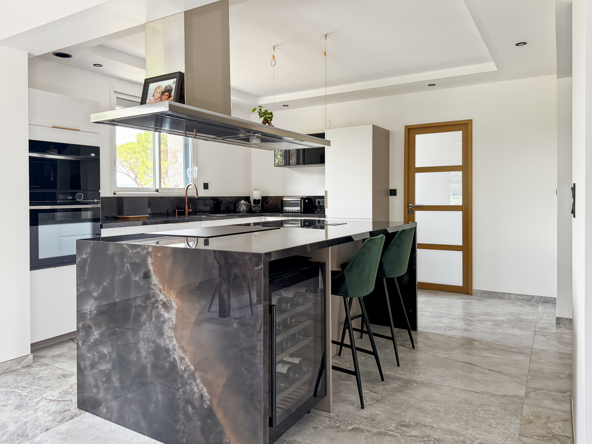 Cuisine moderne avec un îlot central en marbre et des chaises noires, lumineuse et spacieuse.