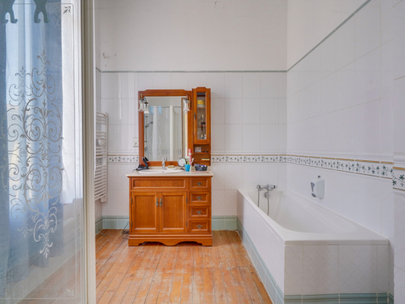Salle de bain avec baignoire et meuble en bois, carrelage blanc et parquet au sol.