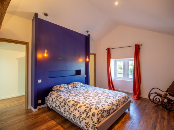 Chambre d'une maison rénovée avec un lit double, murs violets et fenêtre lumineuse.