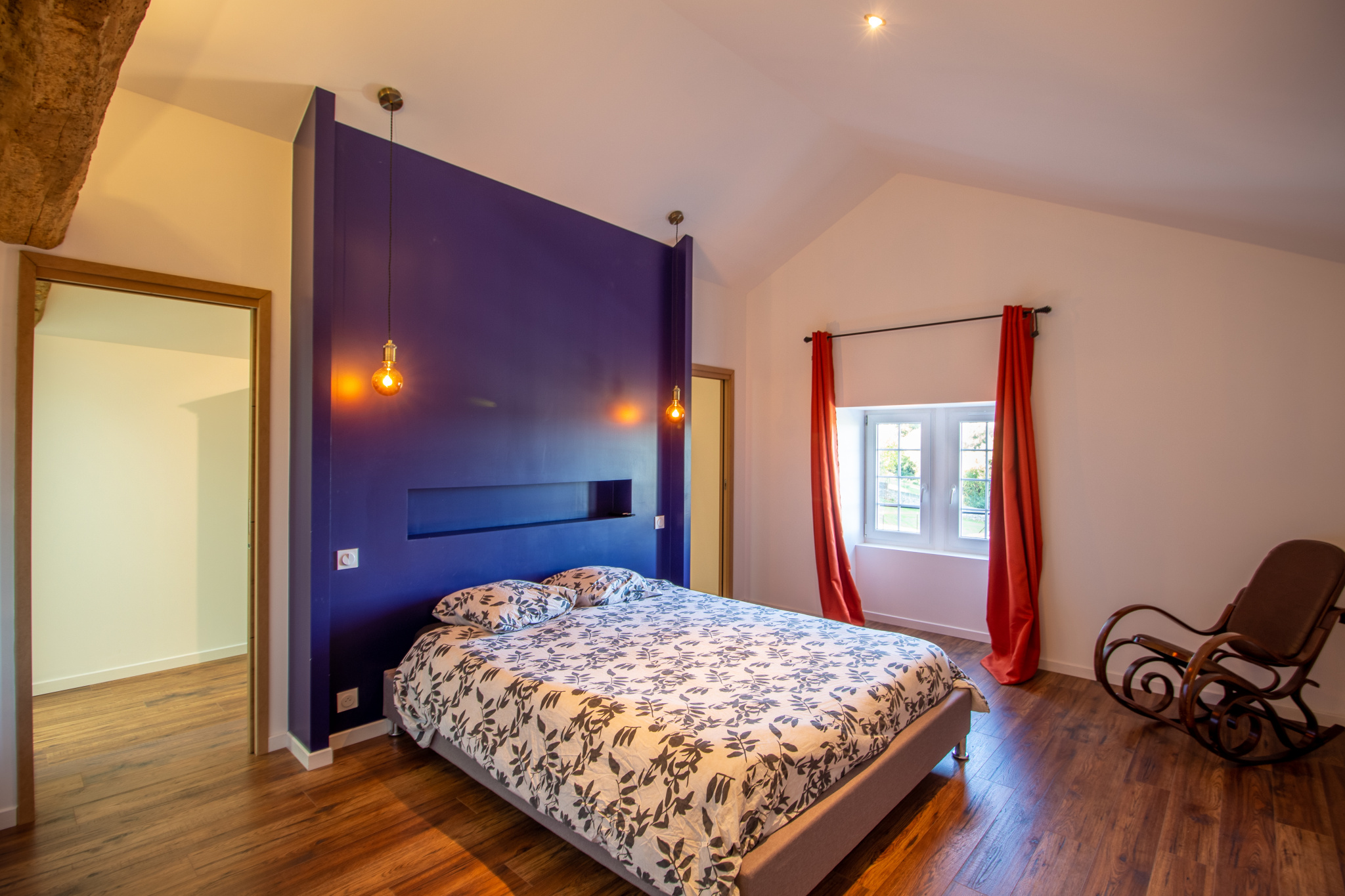 Chambre d'une maison rénovée avec un lit double, murs violets et fenêtre lumineuse.