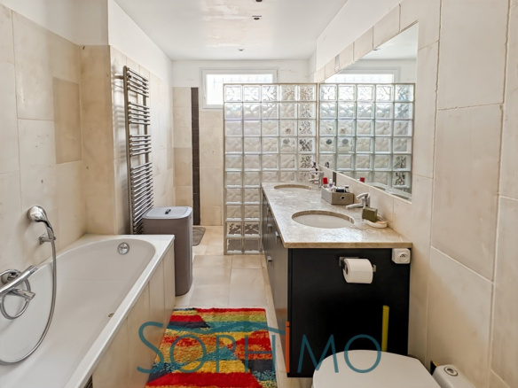 Salle de bain moderne avec baignoire et double vasque, carrelage clair et mur en verre.
