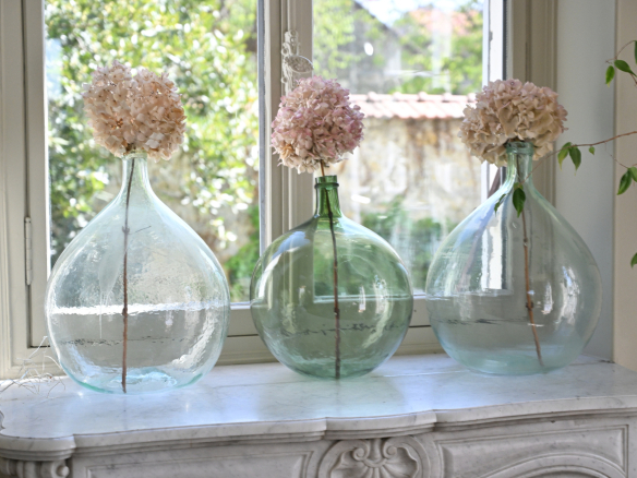 Trois grandes bouteilles en verre avec des fleurs séchées sur une table près d'une fenêtre.