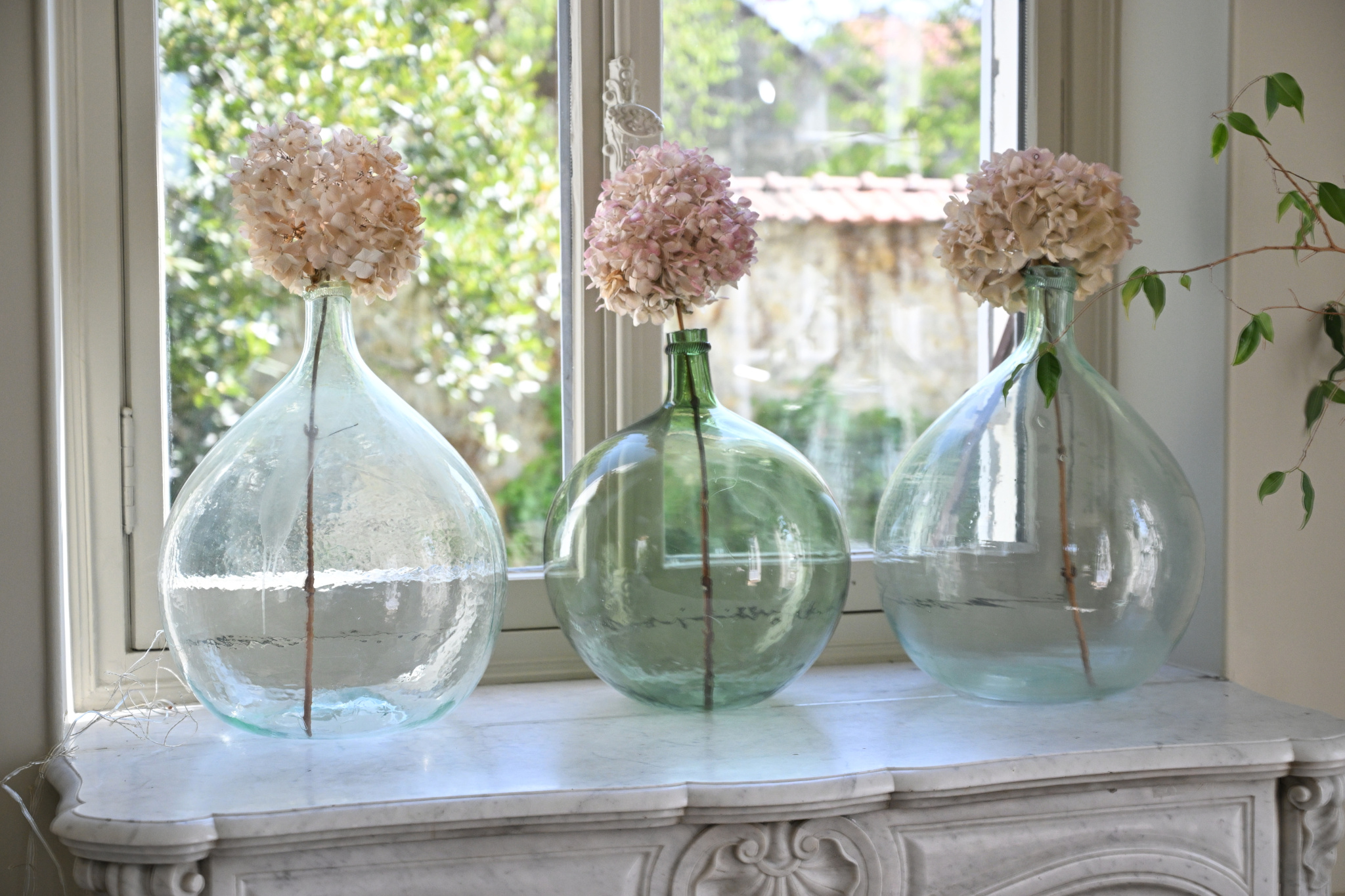 Bouteilles en verre décoratives Trois grandes bouteilles en verre avec des fleurs séchées sur une table près d'une fenêtre.