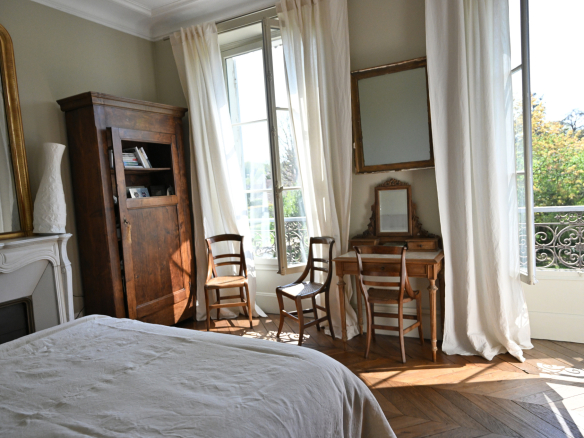 Intérieur d'une chambre avec un lit, des chaises en bois et une commode, fenêtres ouvertes sur un jardin.