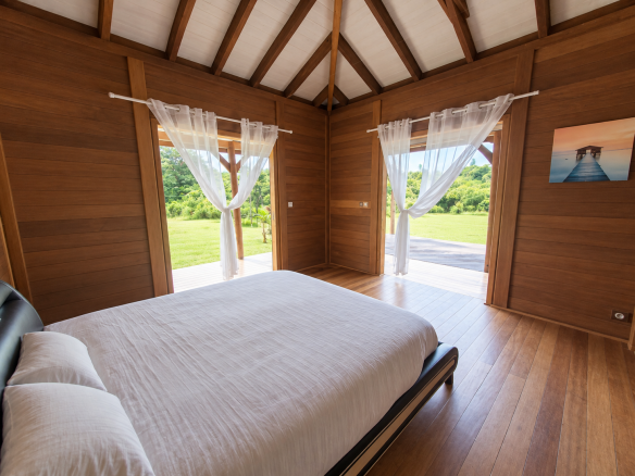 Intérieur d'une maison en bois avec lit et grandes fenêtres donnant sur un jardin verdoyant.