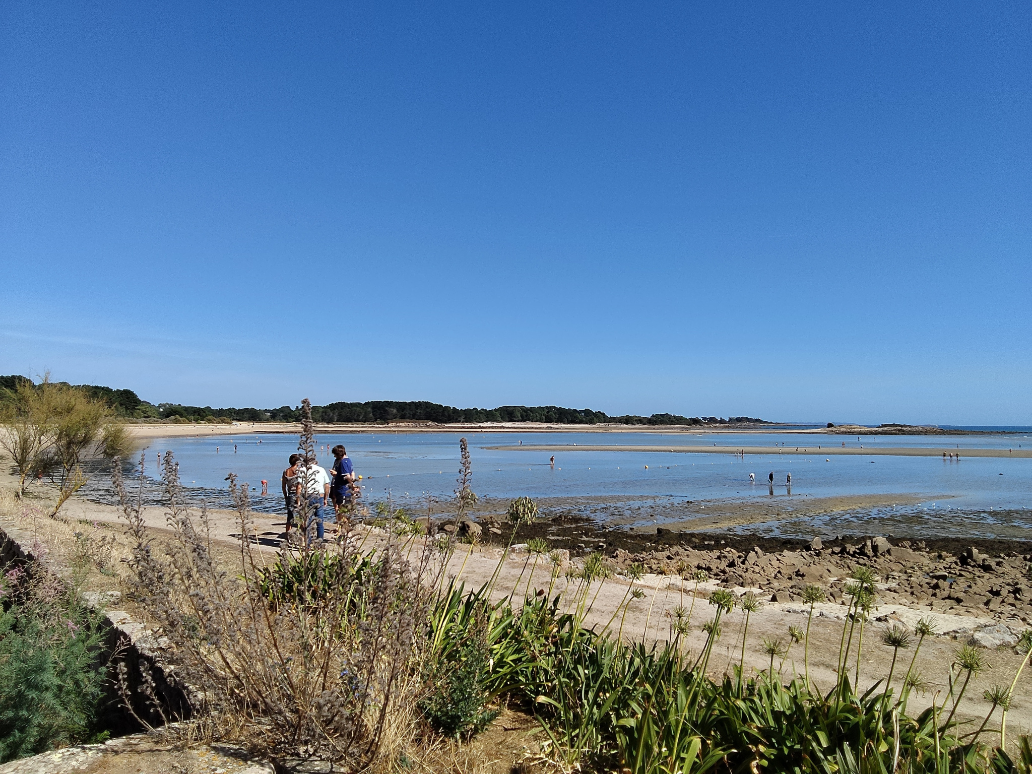 Two people walking on a beach at low tide in Carnac, with a view of the sea.