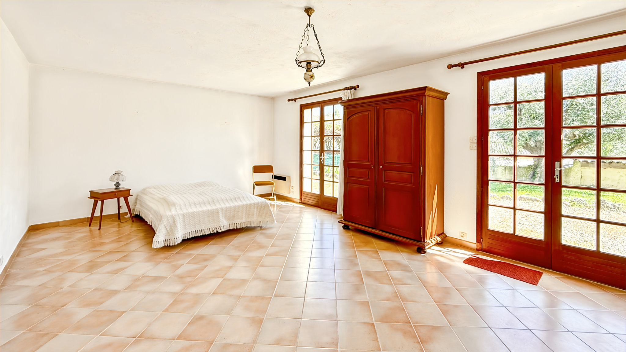Interior of a spacious bedroom with a bed, a wooden piece of furniture and windows overlooking the outside.