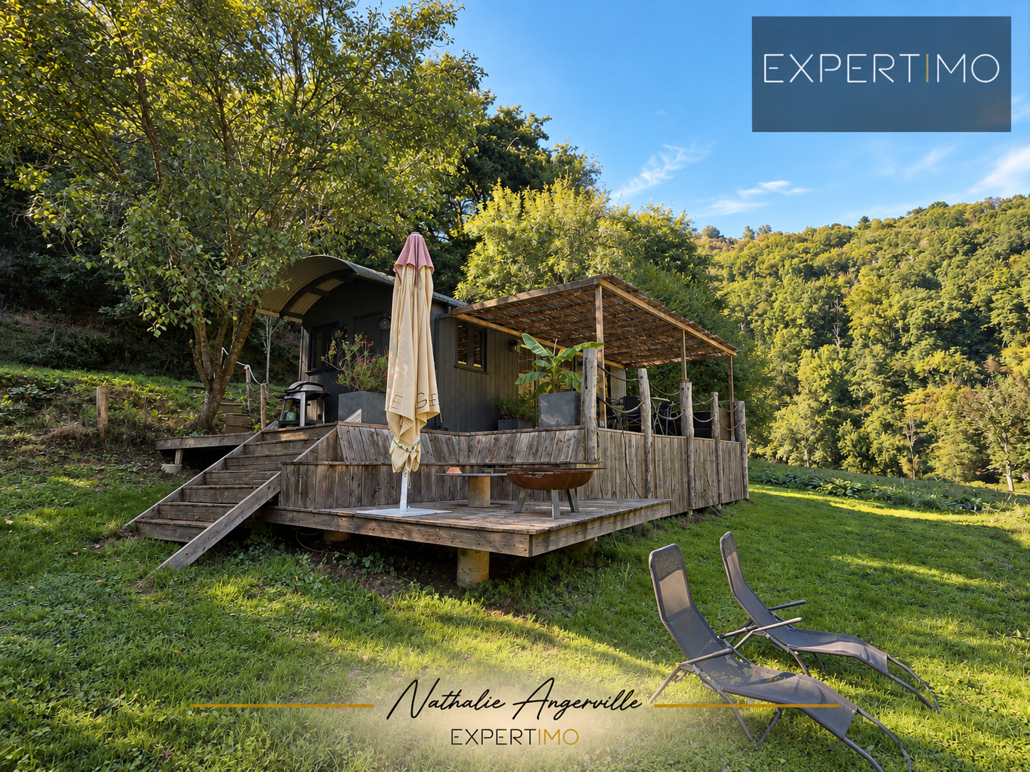 Terrasse d’un ancien moulin réhabilité Terrasse en bois avec parasol et chaises sur un terrain verdoyant, entourée d'arbres.