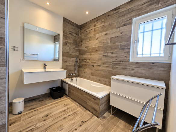 Salle de bain moderne avec baignoire, meuble vasque et miroir, murs en bois et carrelage clair.