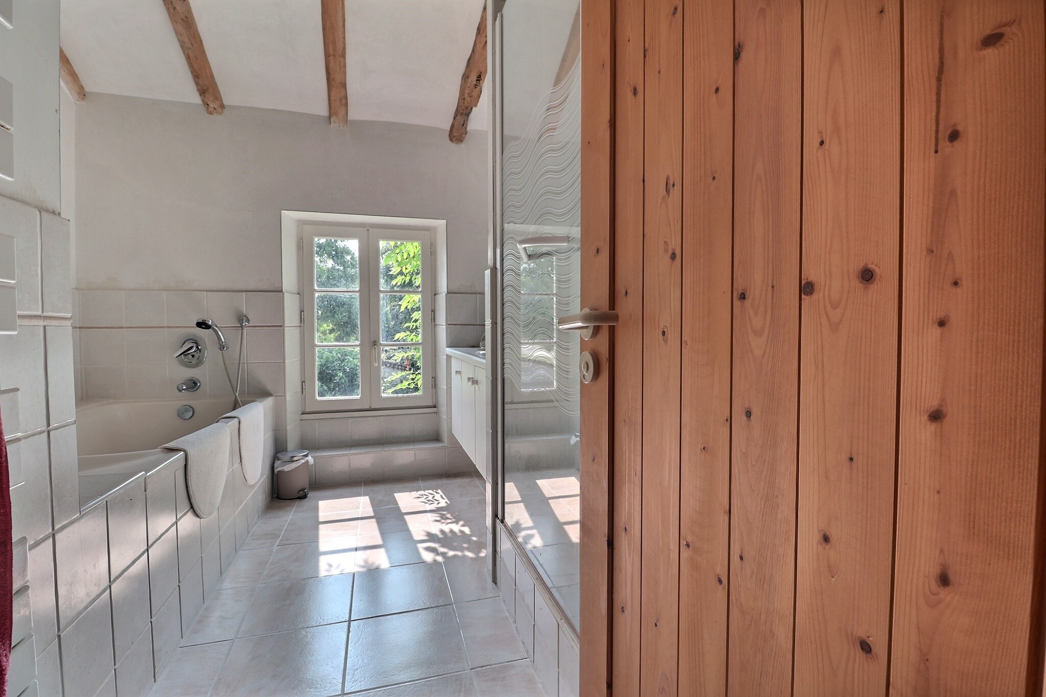 Salle de bain de la propriété à Villeneuve-sur-Lot Salle de bain lumineuse avec baignoire et fenêtre, murs carrelés et plafond à poutres apparentes.