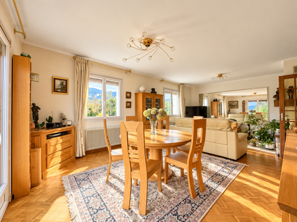 Intérieur d'une maison avec salle à manger lumineuse et salon, fenêtres donnant sur un paysage extérieur.