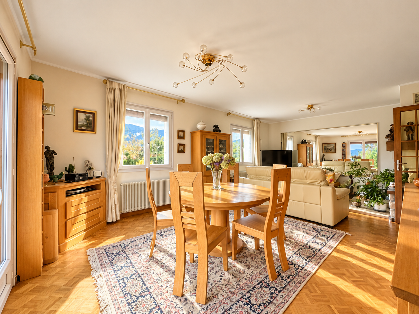 Intérieur d'une maison avec salle à manger lumineuse et salon, fenêtres donnant sur un paysage extérieur.