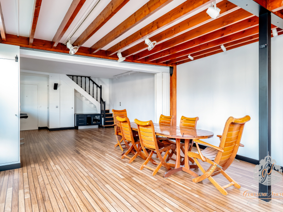 Intérieur d'un duplex avec table en bois et chaises, poutres apparentes, et escalier.