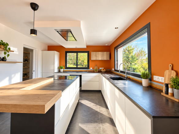 Cuisine moderne avec îlot central, murs orange et fenêtres offrant une vue sur l'extérieur.