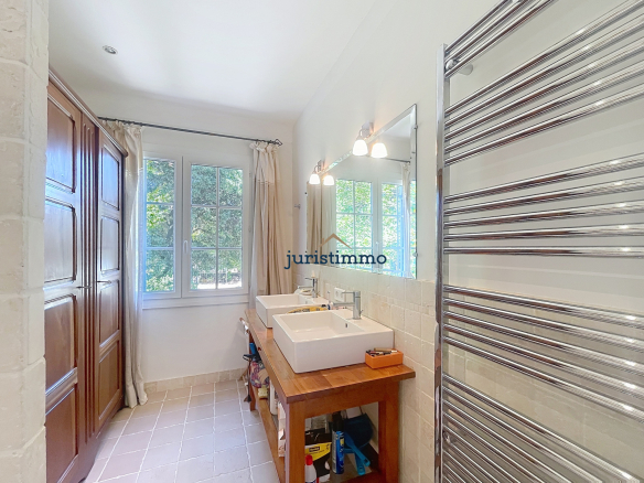 Salle de bain lumineuse avec deux lavabos et un grand miroir, fenêtres donnant sur un jardin.