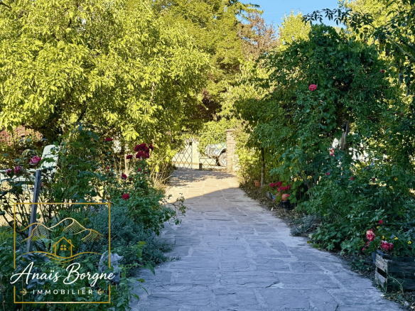 Chemin menant à un manoir dans un parc arboré avec des fleurs et des arbres.