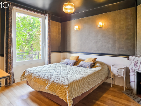Chambre d'une meulière à Arpajon avec lit, cheminée en marbre et fenêtres donnant sur l'extérieur.