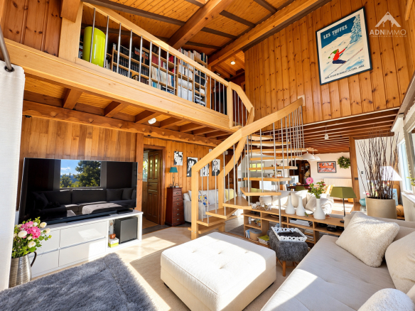 Intérieur d'un chalet en bois avec un salon lumineux, un escalier en bois et des étagères.