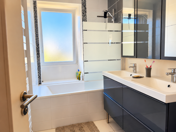 Salle de bain moderne avec baignoire et meuble double vasque, lumière naturelle par la fenêtre.