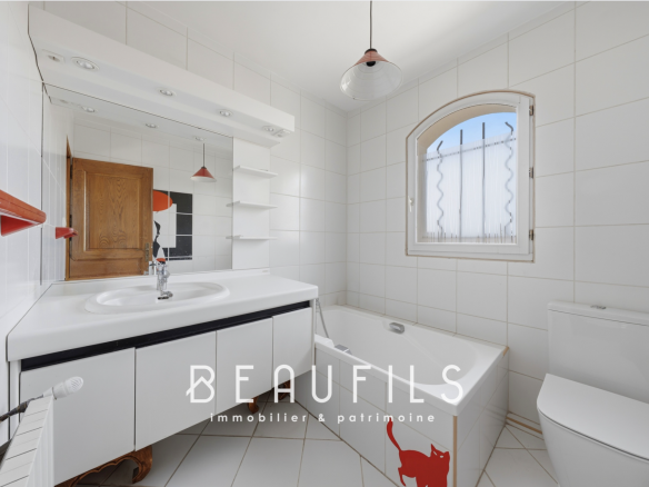 Salle de bain avec baignoire, lavabo et toilettes, carrelage blanc, miroir et fenêtre.