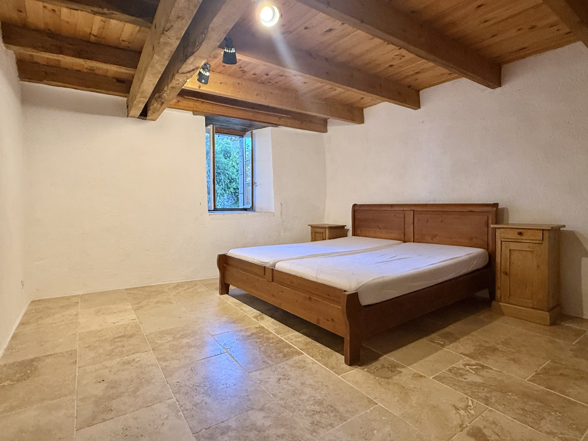Chambre spacieuse avec lit en bois et murs en pierre, éclairée par une fenêtre.