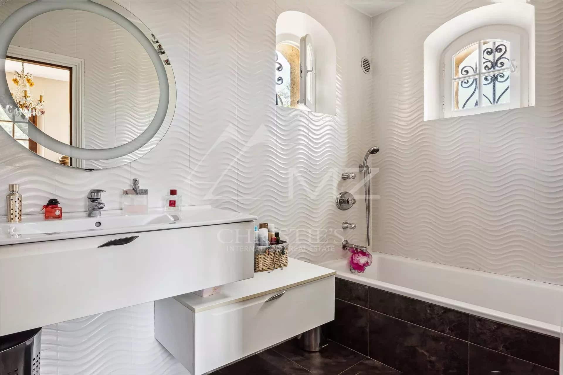 Salle de bain moderne avec baignoire et lavabo, murs ondulés, fenêtre avec grille décorative.