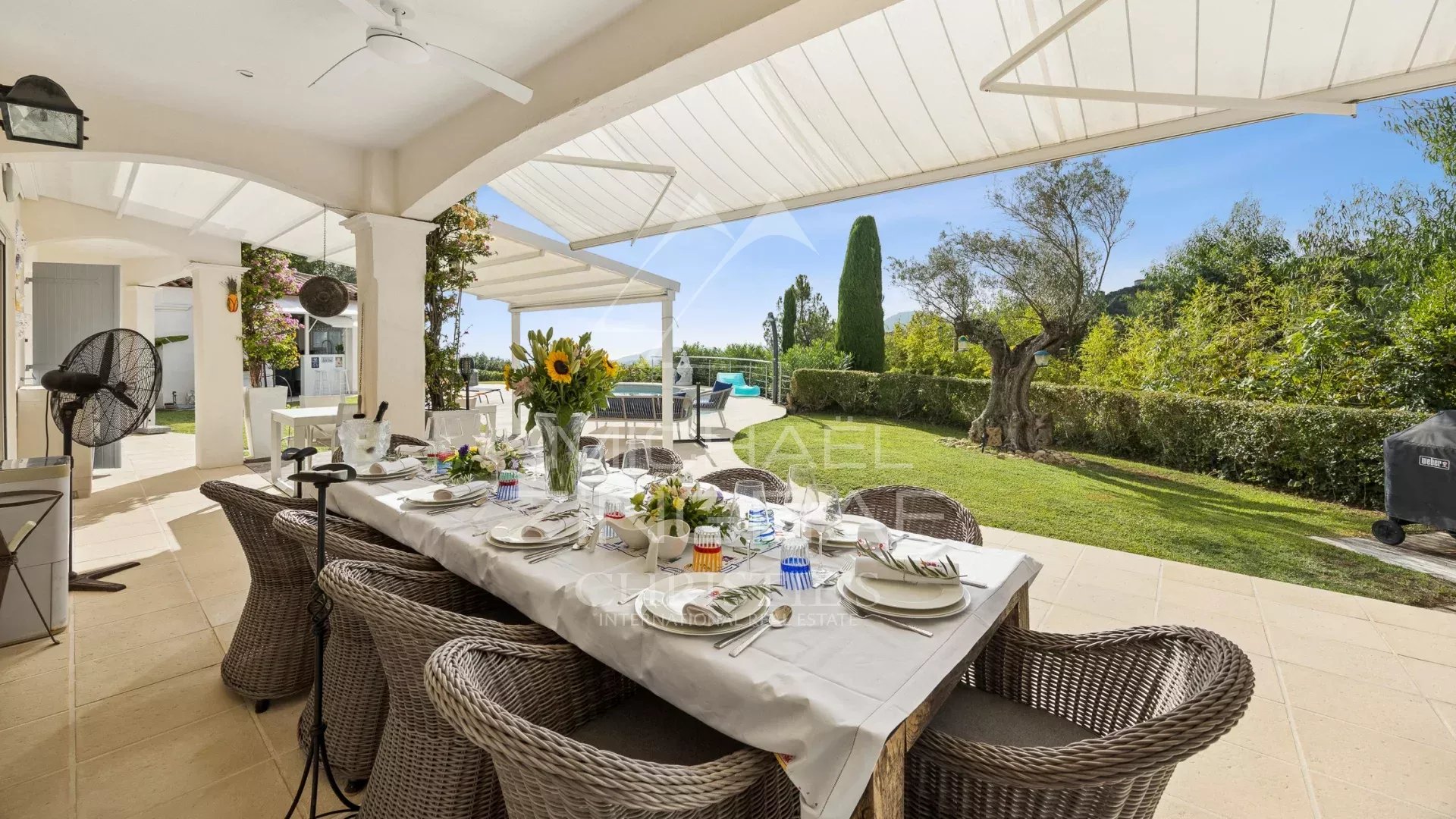 Terrasse d'une propriété avec table dressée et vue sur un jardin verdoyant et la mer.
