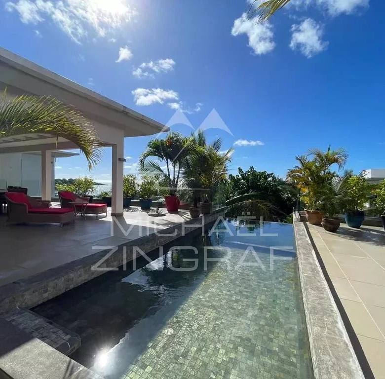 Rooftop poolside seating area with red loungers under a covered patio and potted palm plants against a bright blue sky.