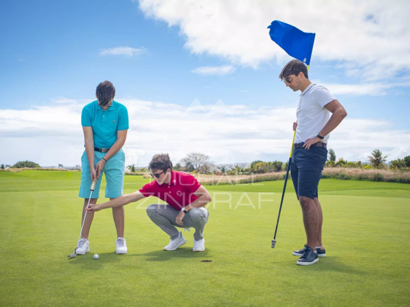Three golfers on a sunny course: one preparing to putt, another crouching and pointing at the ball, and a third standing with a flag on the green in the background.