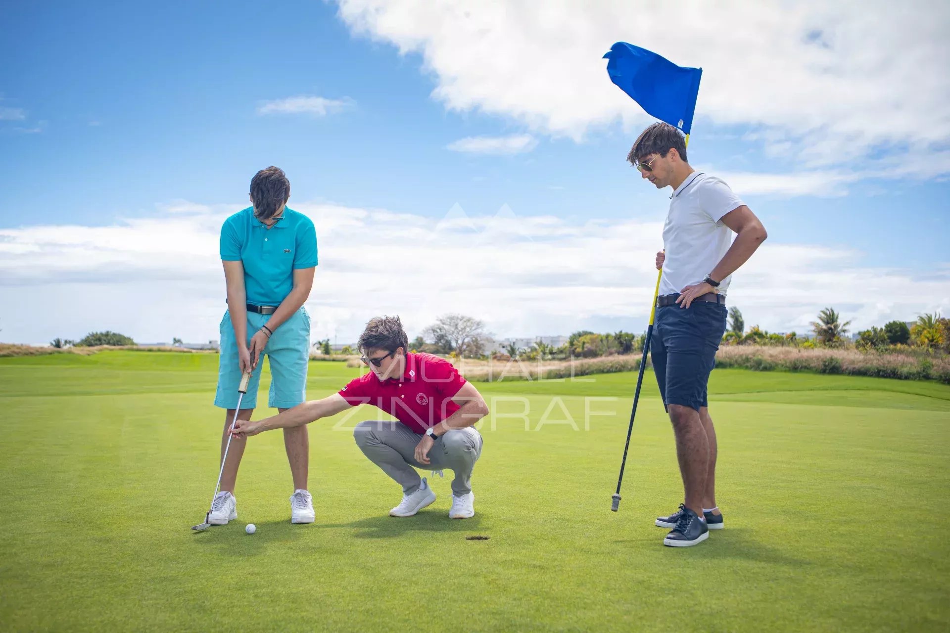 Three golfers on a sunny course: one preparing to putt, another crouching and pointing at the ball, and a third standing with a flag on the green in the background.