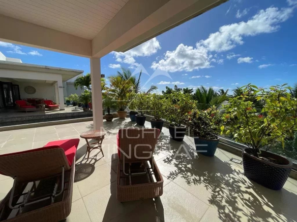 Sunny rooftop terrace with wicker lounge chairs and red cushions, potted plants along the railing, and an ocean view under a blue sky.