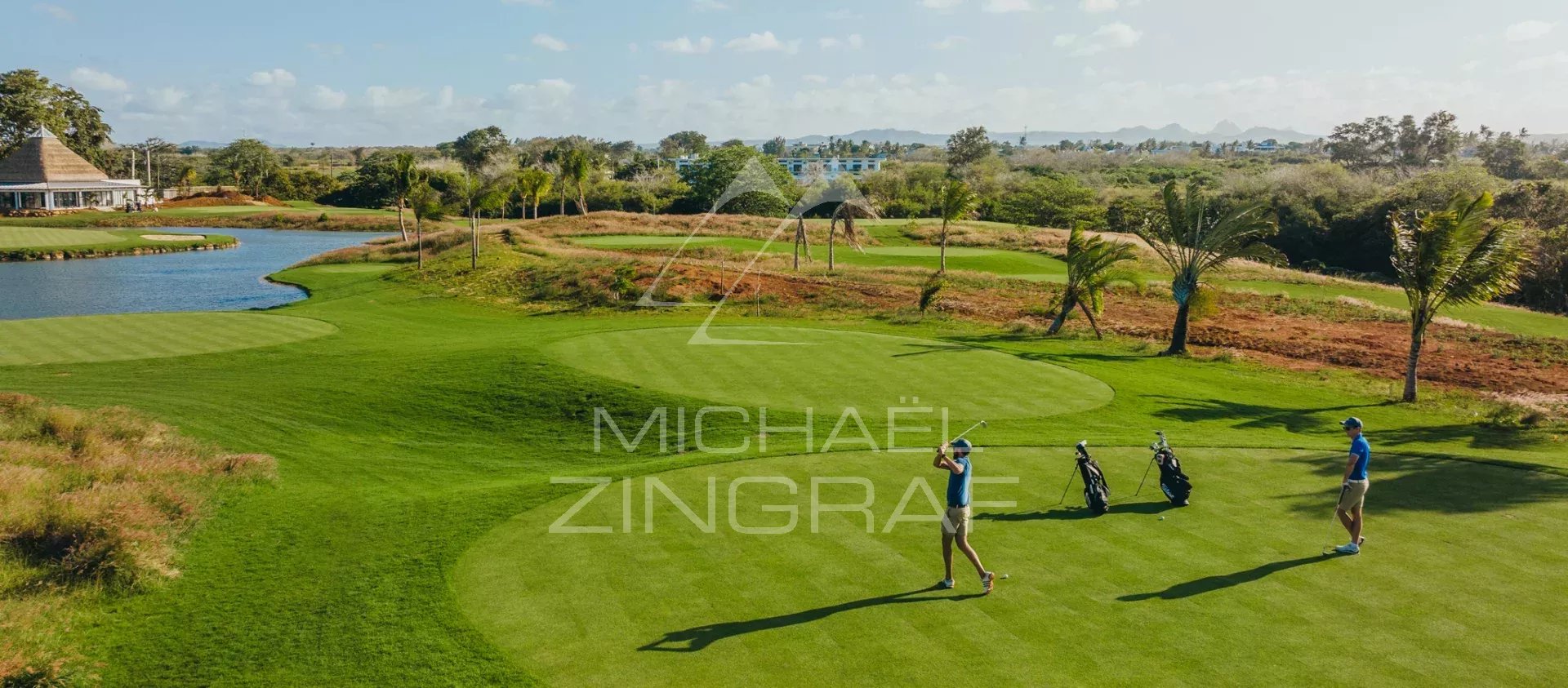 Two golfers on a sunny golf course tee, with golf bags nearby and a water hazard to the left.