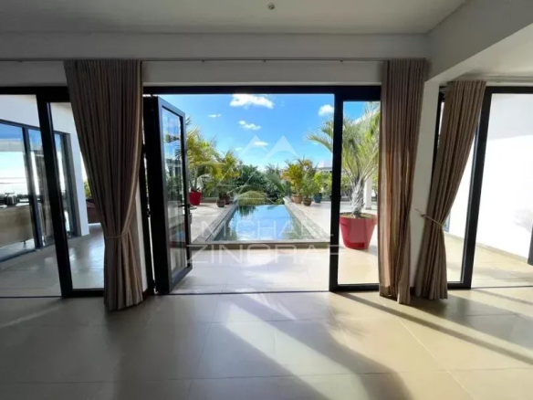 Modern living area with floor-to-ceiling glass doors open to a rectangular pool, potted palms, and a sunny blue sky.
