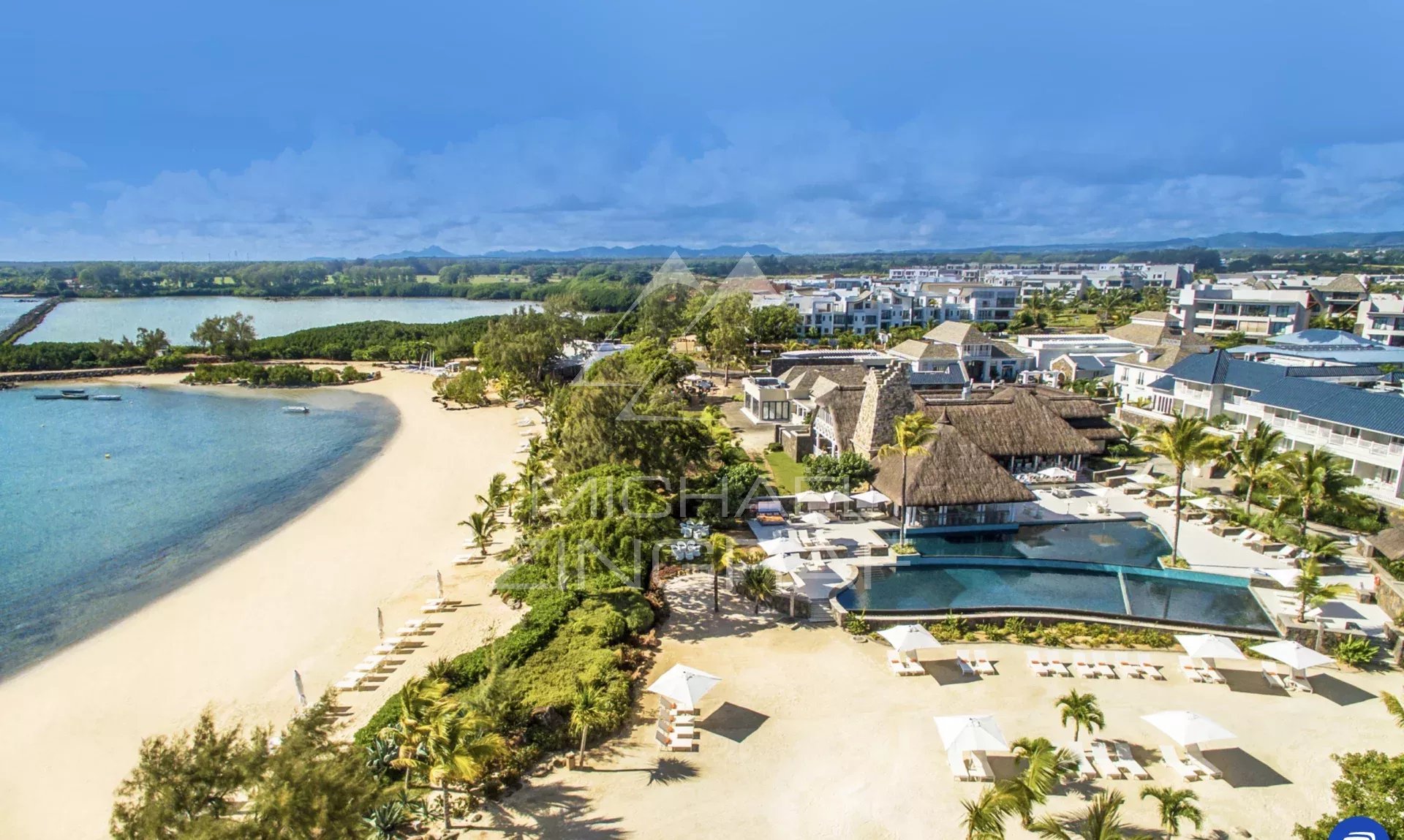 A tropical beachfront resort with a sandy shore, turquoise water, palm trees, and white sun loungers.