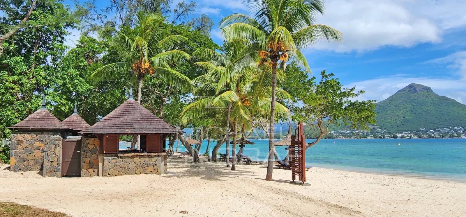 Tropical beach scene with stone cabanas, palm trees, and calm turquoise water, mountain in the distance