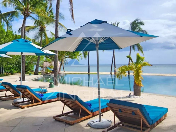Poolside lounge area with blue-cushioned chairs and blue umbrellas overlooking a calm ocean and sandy beach, tropical trees in the background.