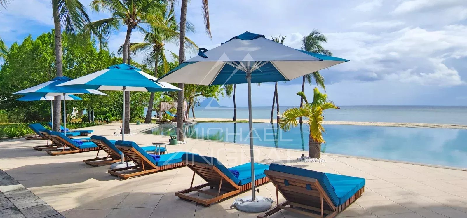 Poolside lounge area with blue-cushioned chairs and blue umbrellas overlooking a calm ocean and sandy beach, tropical trees in the background.