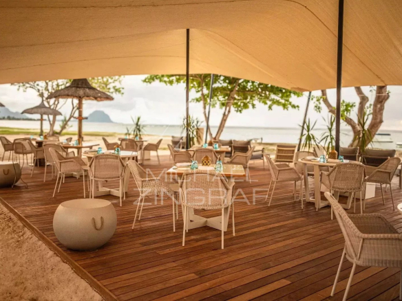 Beachside outdoor cafe with wicker chairs and white tables on a wooden deck, umbrellas, and the sea in the background.