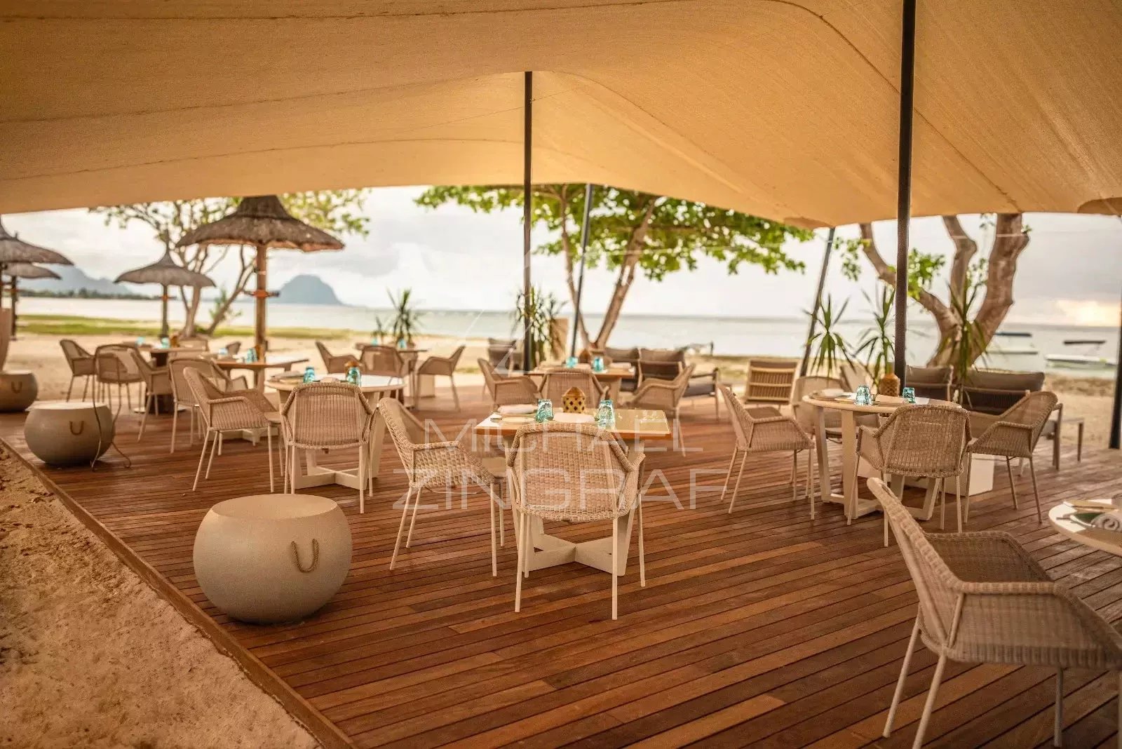 Beachside outdoor cafe with wicker chairs and white tables on a wooden deck, umbrellas, and the sea in the background.