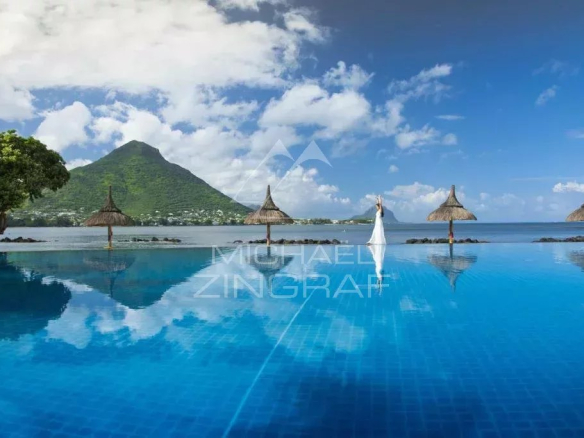 Infinity pool reflecting a bright sky, with thatched umbrellas and a person in a white dress at the edge, mountains in the background.
