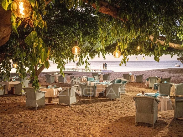 Beachside restaurant setup on the sand with white-tablecloth tables and wicker chairs under a leafy tree, lanterns casting warm light over the scene by the ocean.