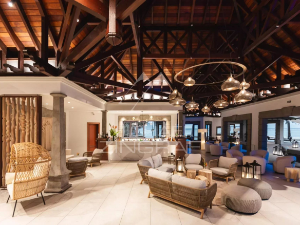 Luxurious open-air lounge with wicker sofas, round ottomans, and a central bar under dark wooden beams; view of the seaside through glass walls.