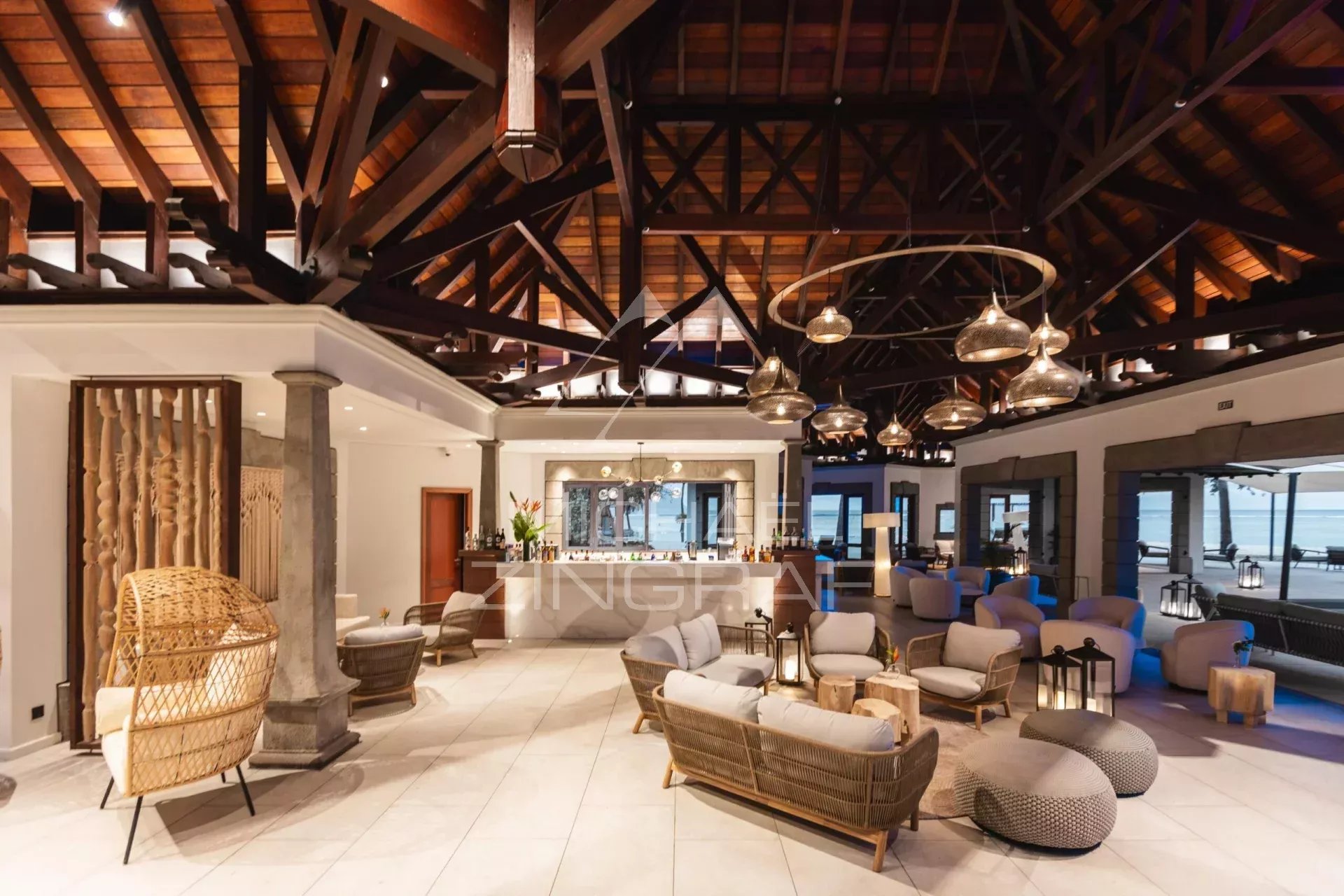 Luxurious open-air lounge with wicker sofas, round ottomans, and a central bar under dark wooden beams; view of the seaside through glass walls.