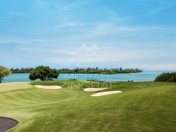Golf course green with two sand bunkers, overlooking turquoise water and trees under a clear blue sky.