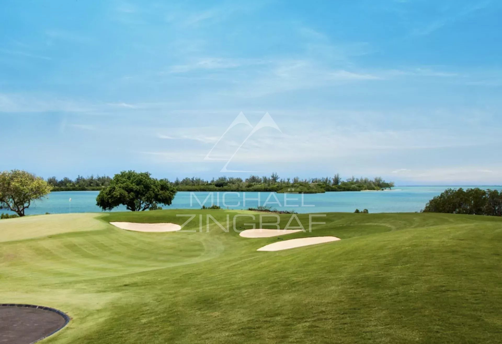 Golf course green with two sand bunkers, overlooking turquoise water and trees under a clear blue sky.