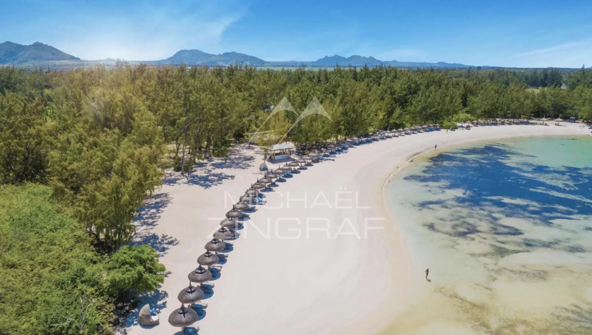 Aerial view of a crescent white-sand beach lined with thatched umbrellas and a forest backdrop, turquoise water at the shore.