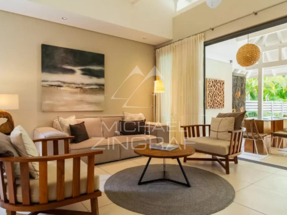Bright living room with a beige sofa, wooden armchairs, round coffee table, and abstract wall art, opening to a patio with outdoor seating through sliding glass doors.