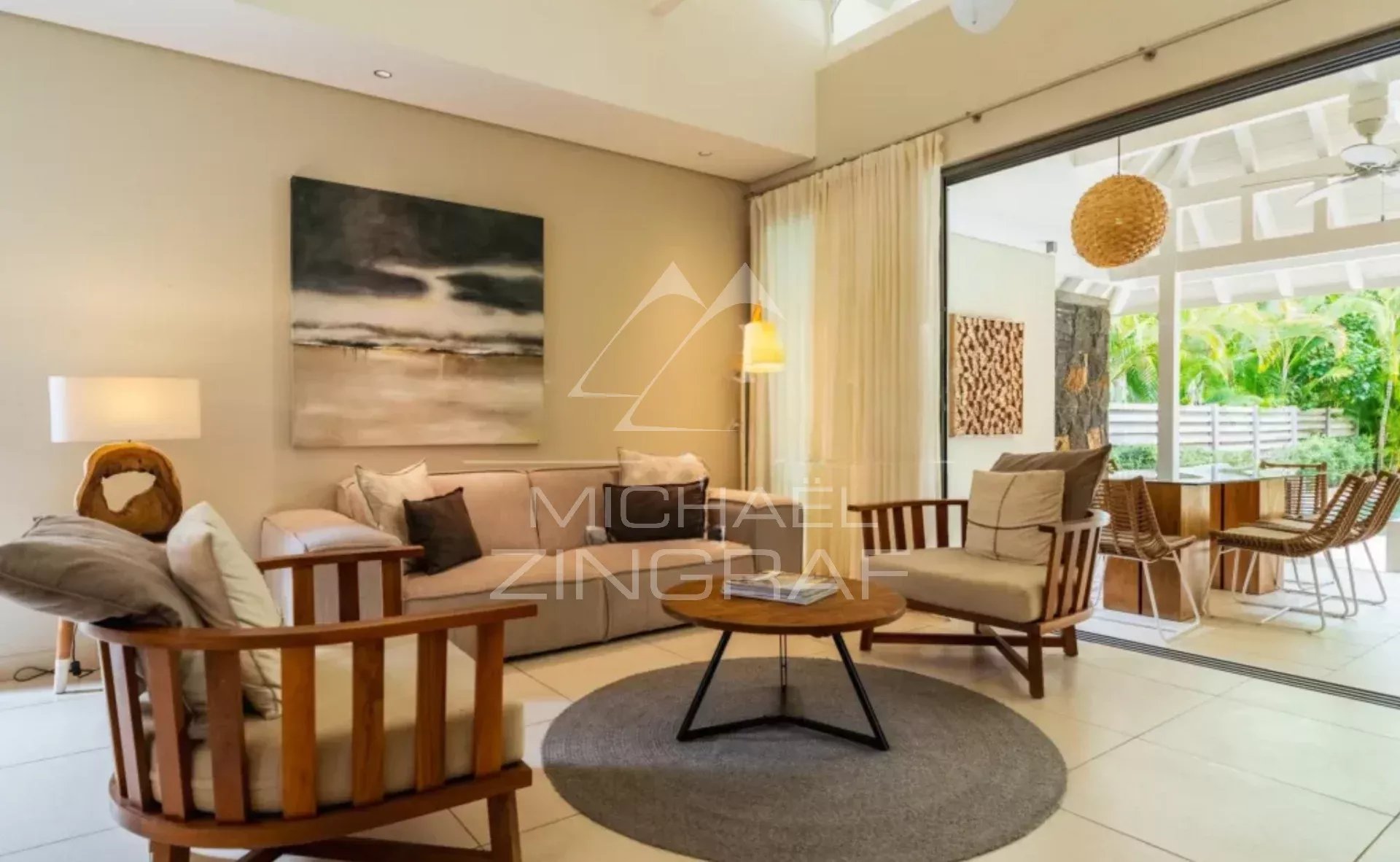 Bright living room with a beige sofa, wooden armchairs, round coffee table, and abstract wall art, opening to a patio with outdoor seating through sliding glass doors.